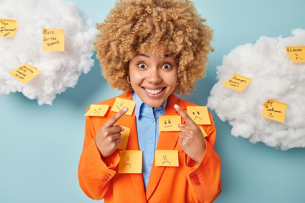 Positive female entrepreneur dressed in formal orange jacket\
points at toothy smile covered with reminding stickers being in\
good mood has various tasks to do isolated on blue background\
clouds above