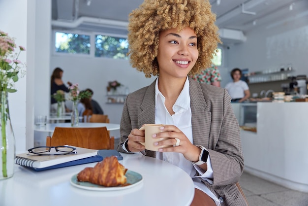 Positive female entrepreneur with curly hair dressed in formal\
outfit holds mug of coffee poses at table with notepads being in\
cafeteria against blurred background looks aside people and\
lifestyle