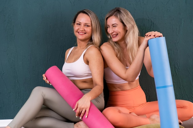 Positive female friends in sportswear holding colorful mats sitting on floor and looking in camera