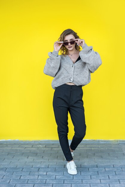 Positive  female model  posing in gray knitted sweater in sunglasses near yellow wall, outdoor