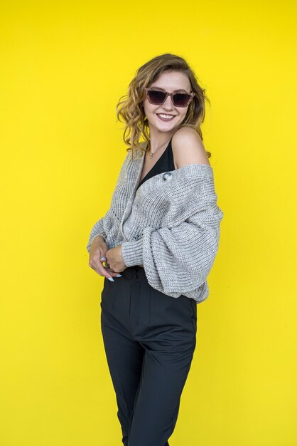 Positive  female model  posing in gray knitted sweater in sunglasses near yellow wall, outdoor