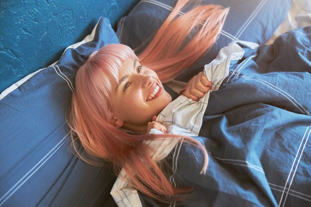 Positive female model with bright pink hair lies in large bed in early morning