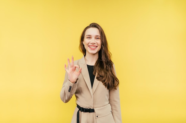 Positive female office worker in suit stands on yellow background with smile on face and shows