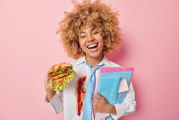 Positive female student eats delicious hamburger carries folders with sticker has tasty snack during break at university wears white formal shirt smeared with ketchup blue tie isolated over pink wall