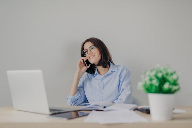 Positive female student in round glasses prepares for exam or makes diploma paper happy to hear her best friend via cell phone share her success at university and future plans after graduating