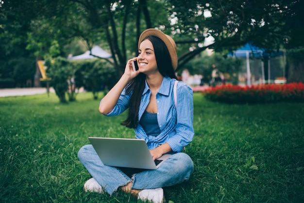 Positive female teenager enjoying cellphone conversation during\
remote work on laptop in city park