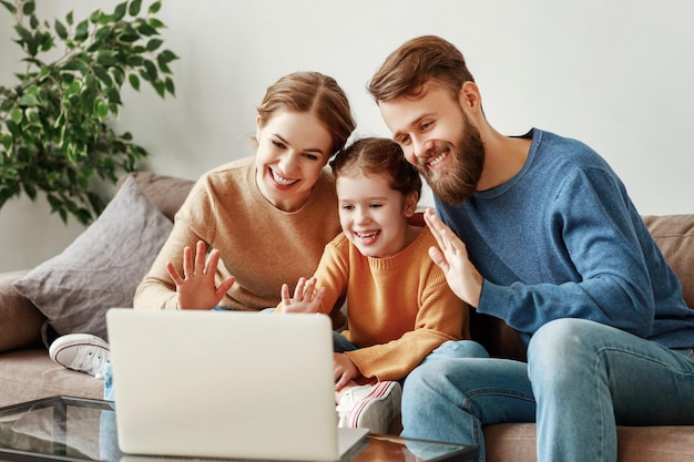 Photo positive friendly young parents with smiling little daughter sitting on sofa together answering video call on laptop and waving hand in greeting while relaxing at home on weekend