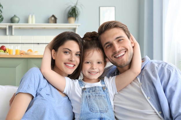 Positive friendly young parents with smiling little daughter sitting on sofa together while relaxing at home on weekend.