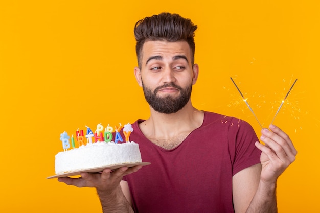 Positive funny young asian guy with a cap and a burning candle and a cake in his hands posing on a yellow wall. Anniversary and birthday concept.