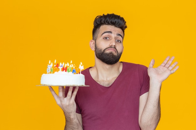 Positive funny young guy with a cap and a burning candle and a homemade cake in his hands posing on