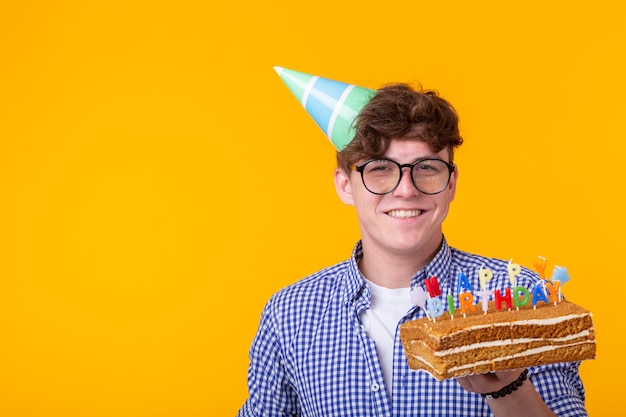 Positive funny young guy with a cap and a homemade cake in his hands posing on a yellow wall