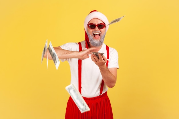 Positive generous grey bearded man in santa claus hat sunglasses and pants with suspenders throwing dollar cash into air celebrating winter holidays indoor studio shot isolated on yellow background