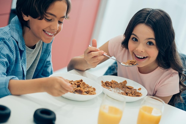 Positive girl and boy sitting with bowls of tasty cornflakes and smiling while eating them