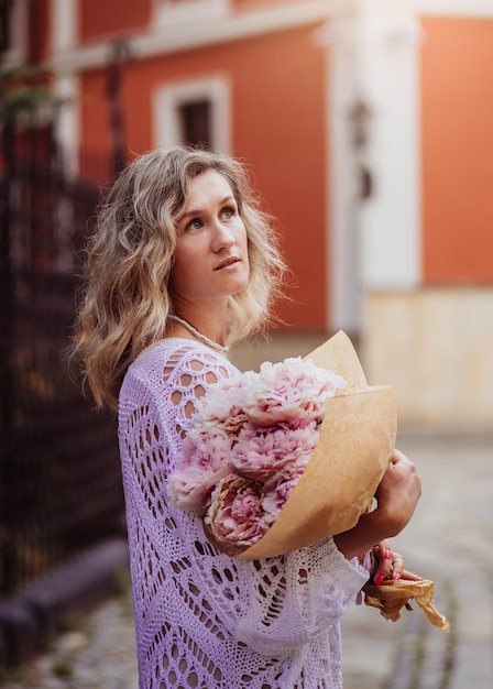 Positive girl hold flowers in the city center Close up shot of amazing blonde haired lady with natural make up look happy walking outdoors hold pink bouquet