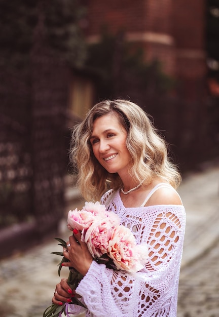 Positive girl hold flowers in the city center Close up shot of amazing blonde haired lady with natural make up look happy walking outdoors hold pink bouquet