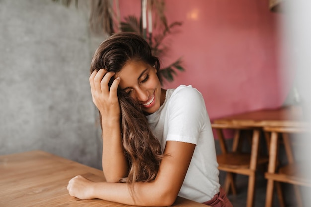 Фото Позитивная девушка в белой футболке сидит за деревянным столом в кафе. снимок кудрявой женщины в отличном настроении.