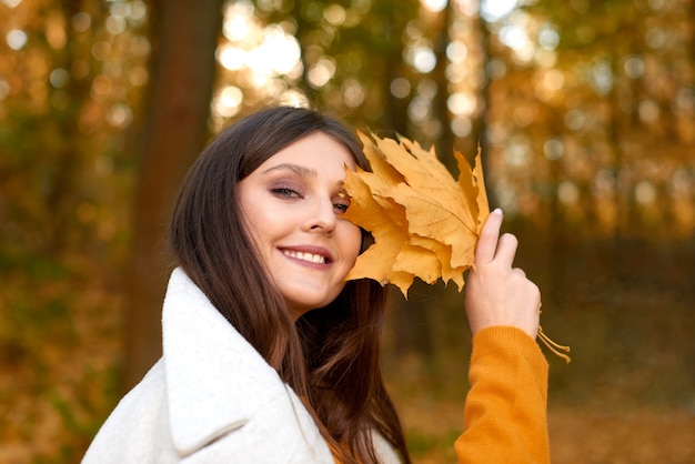 Positive girl laughs and covers her face with bouquet of dry fallen leaves in park or forest in fall