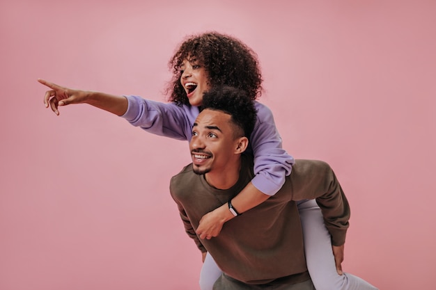 Positive girl in purple outfit sitting on her boyfriends back and pointing to left