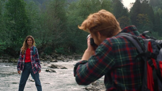 Positive girl on river bank posing for male photographer redhead guy taking photos of female model in mountains smiling woman showing peace sign with hands outdoor