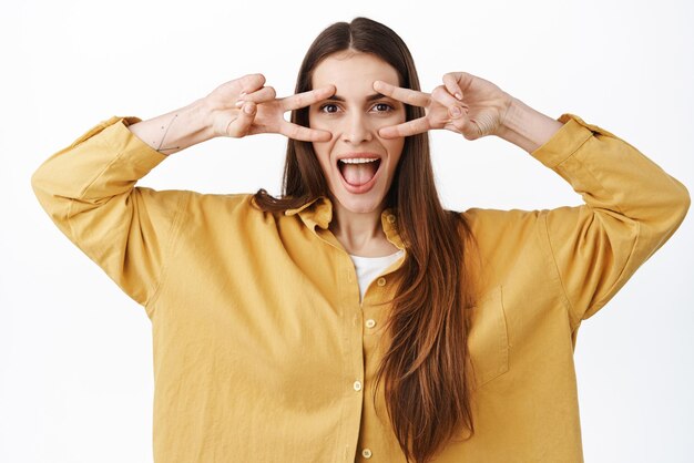 Positive girl shows peace signs and smiles say hey looking through vsigns with happy expression standing against white background