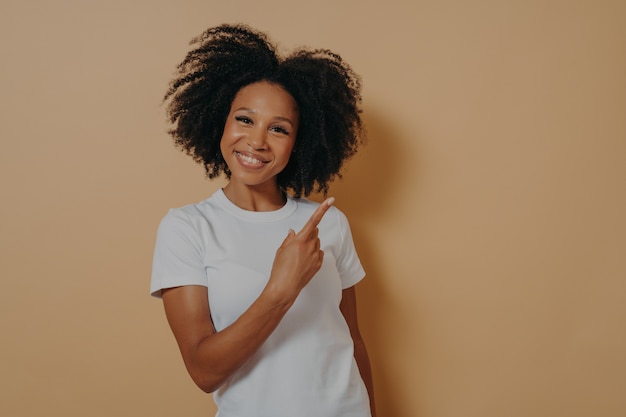 Positive good looking african female model in white shirt indicating with forefinger at blank copy space and smiling, pointing at right upper corner. Advertisement and promotion concept