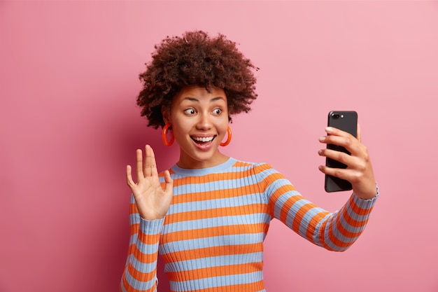 Positive good looking Afro American woman talks selfie and waves hand at smartphone camera smiles happily wears long sleeved striped jumper isolated over pink wall