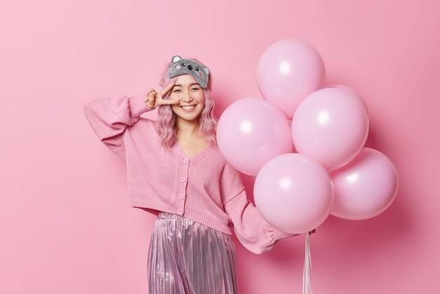 Positive good looking pink haired woman enjoys festive event comes to congratulate friend with birthday makes peace gesture over eye poses with helium balloons wears jumper skirt and sleepmask