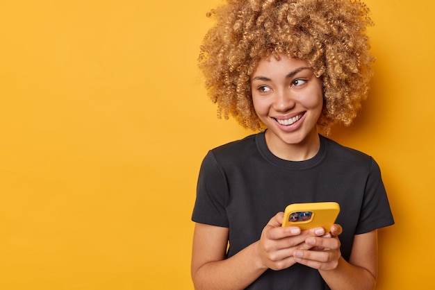 Positive good looking young woman with cheerful smile on face curly hair uses smartphone chats online wears casual black t shirt isolated over yellow background blank space for your advertising