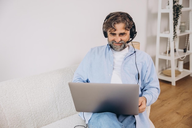 Positive grandfather using laptop computer and headset sitting on couch Senior man working from home have online meeting video chat copy space