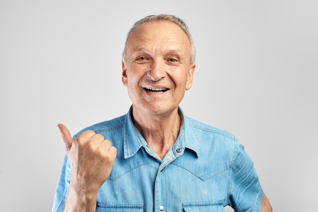 Positive gray-haired senior citizen joyfully smiles, shows a finger to the side on free copy space isolated in white studio