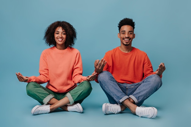 Positive guy and his girlfriend meditate on isolated background Charming curly woman in green jeans and darkskinned man in red sweater smile on blue backdrop