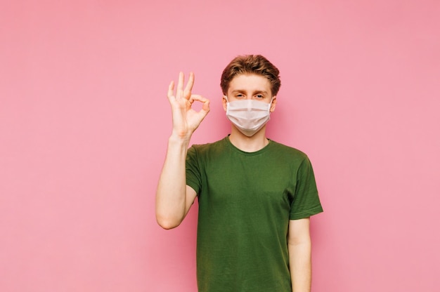 Positive guy in a medical mask isolated on a pink background looks at the camera with a smile and shows a OK gesture Cheerful guy in quarantine wears a mask and shows a quotokayquot gesture