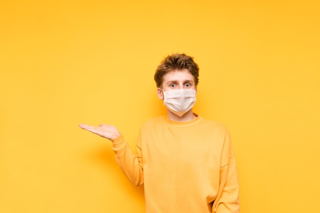 Позитивный парень в оранжевой толстовке и белой медицинской маске стоит на желтом фоне