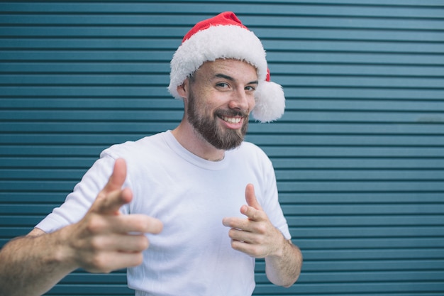 Positive guy with santa hat is smiling. He is pointing on camera. Bearded guy is happy. Isolated on striped 