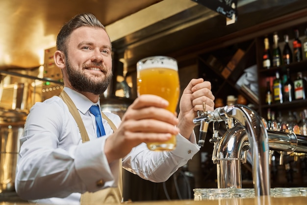 Positive, handsome barman holding cold lager beer glass. Cheerful brewery worker standing at bar counter, smiling. Adult man in white shirt and brown apron looking at camera.