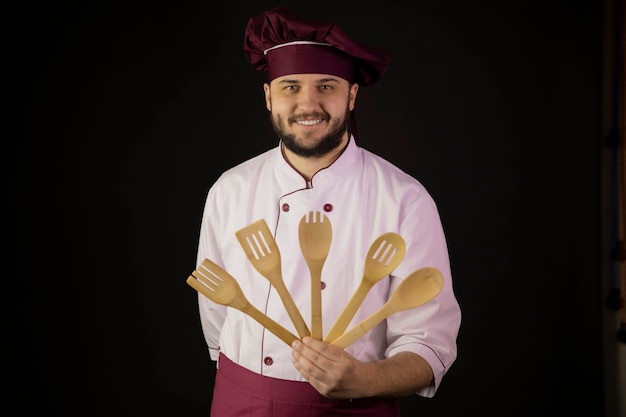 Positive handsome bearded chef man in uniform holds wooden kitchen utencils