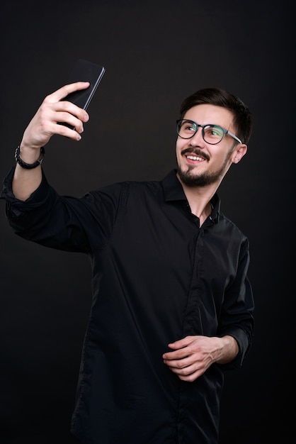 Positive handsome young man in eyeglasses taking selfie in black shirt on smartphone against isolated dark background