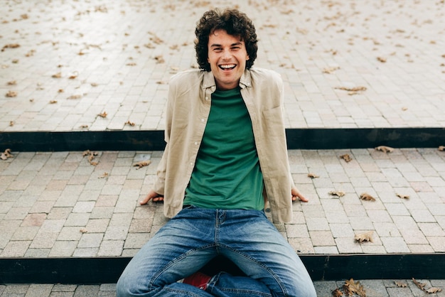 Photo positive handsome young man sitting on the stairs smiling and looking at the camera outdoor portrait of happy smart student male resting outside people and emotion concept