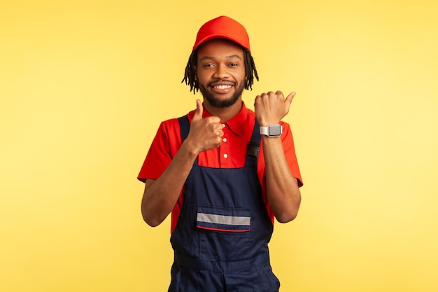 Positive handyman or builder showing smart watch on his hand and thumb up, like gesture, satisfied with excellent delivery service on time. Indoor studio shot isolated on yellow background.