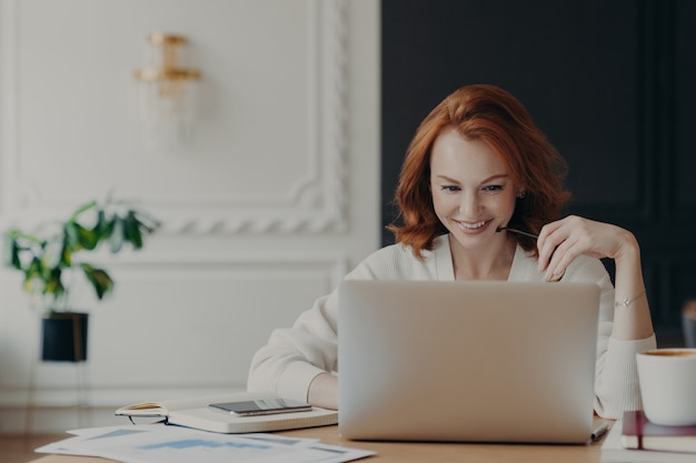 Positive happy female freelancer has busy working day, works distantly from home, sits in front of laptop computer against modern interior, works on creative task, watches webinar for improving skills