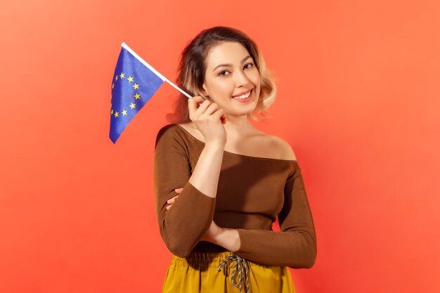 Positive happy woman holding flag of europe union looking at
camera with toothy smile happy of open borders indoor studio shot
isolated on orange background