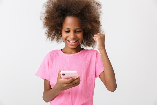 ポジティブな幸せな若いアフリカの女の子の子供が白い壁の上に孤立してポーズをとって、携帯電話を使用して勝者のジェスチャーをします。