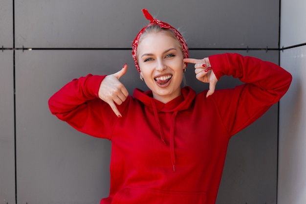 Positive happy young woman with a cute smile in a stylish red sweatshirt with a trendy bandana posing in a city near a gray building on a summer day