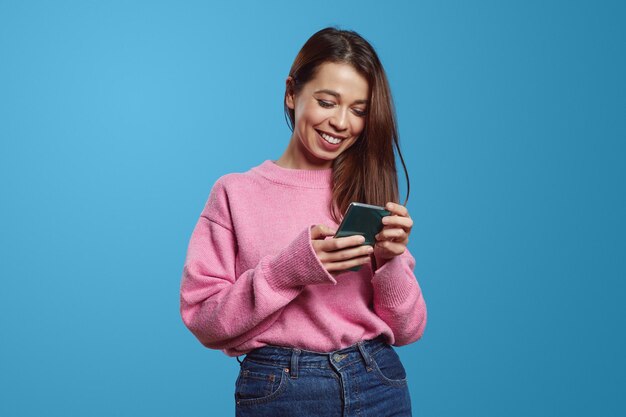 Foto femmina ispanica positiva in maglione rosa che sorride felicemente mentre utilizza lo smartphone contro la parete blu