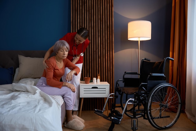 Positive home caregiver helping sad unhappy old woman in bedroom