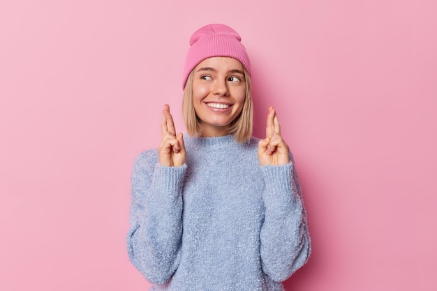 Positive hopeful woman smiles happily keeps fingers crossed makes wish looks away dressed in casual clothes poses against pink background waits for something with excitement Body language concept