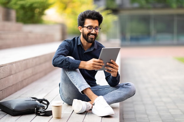 Positive indian guy resting at park after work using tablet