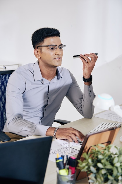 Photo positive indian programmer in glasses working on computer and recording voice message for coworker