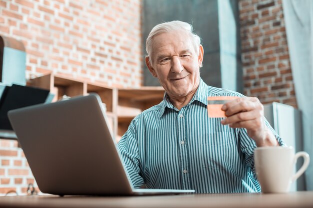 Positive intelligent aged man sitting in front of the computer and looking at the credit card while learning how to use online banking system