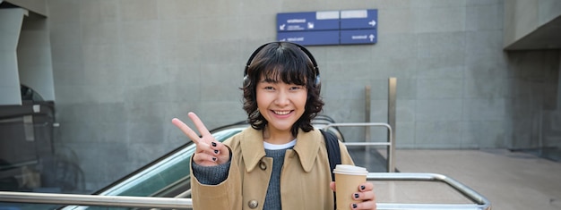 Positive korean girl in headphones drinks coffee to go shows peace vsign stands on street and smiles